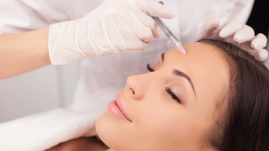 Close up of hands of expert beautician injecting botox in female forehead. The woman closed her eyes with joy. She is gently smiling